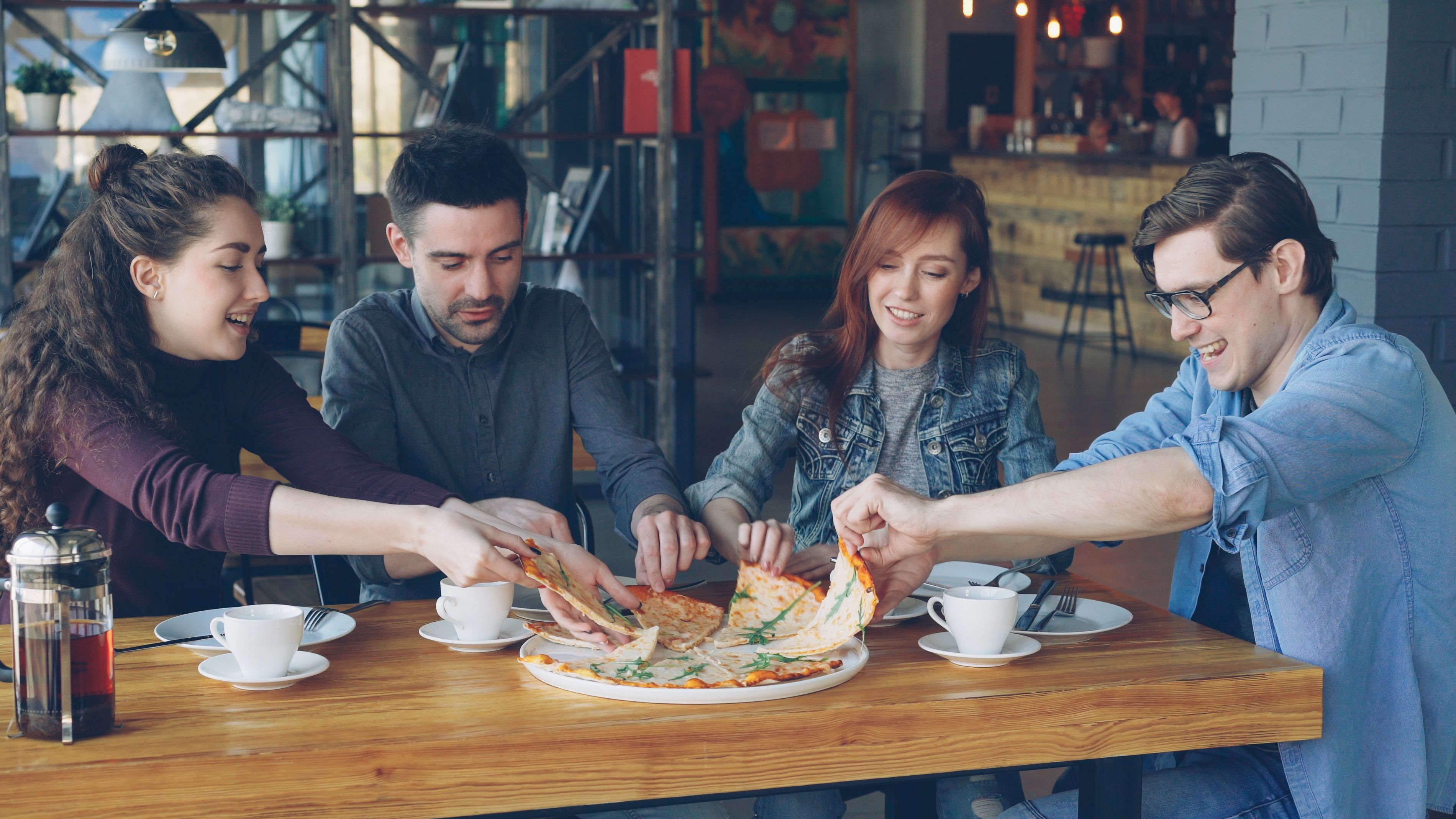 Four friends sitting around a wooden dining table while grabbing slices of pizza together at a lively cafe.